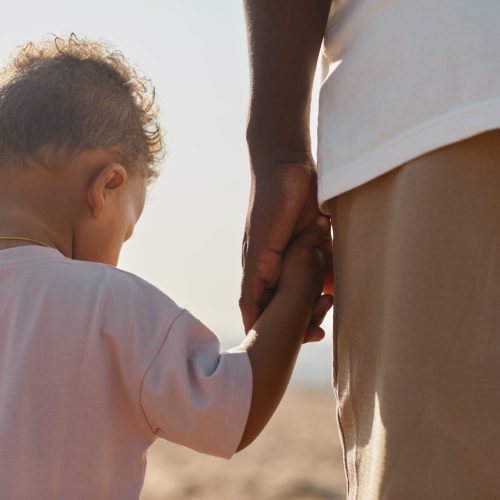 Back view portrait of cute African-American toddler holding hands with dad while enjoying walk on beach in sunlight, copy space
